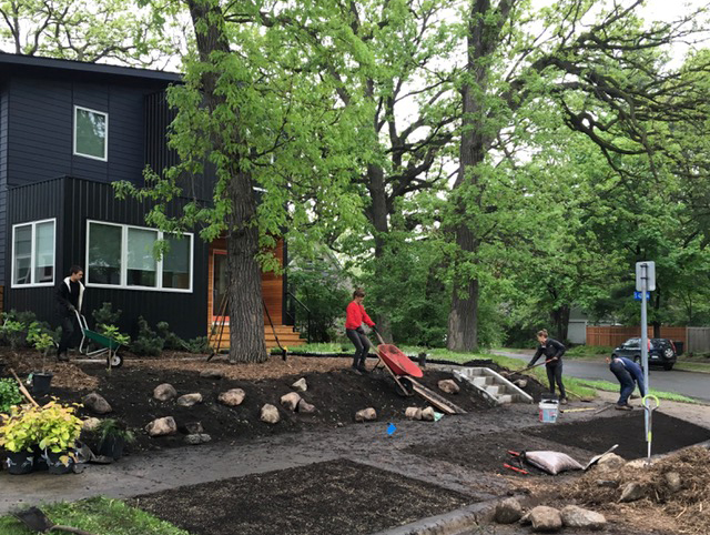 Four landscape crew members installing new boulders and plants into fresh dirt in front of a modern black and natural wood house on an urban street.