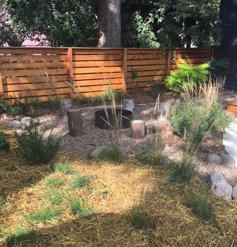 A rustic metal fire pit with wooden stumps for seating surrounding it, in a backyard of a modern black house with a natural wood fence.