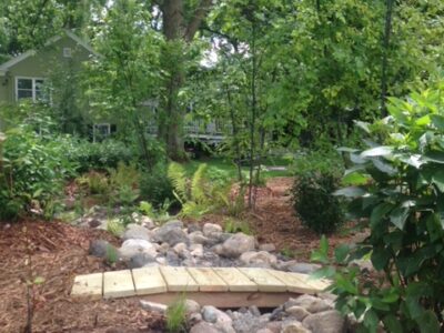 A dry rocky creek bed with a wooden bridge running over it and a light green house in the background.