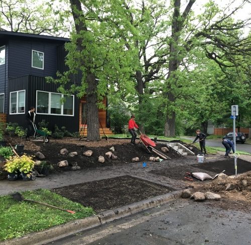 Four landscape crew members installing new boulders and plants into fresh dirt in front of a modern black and natural wood house on an urban street.