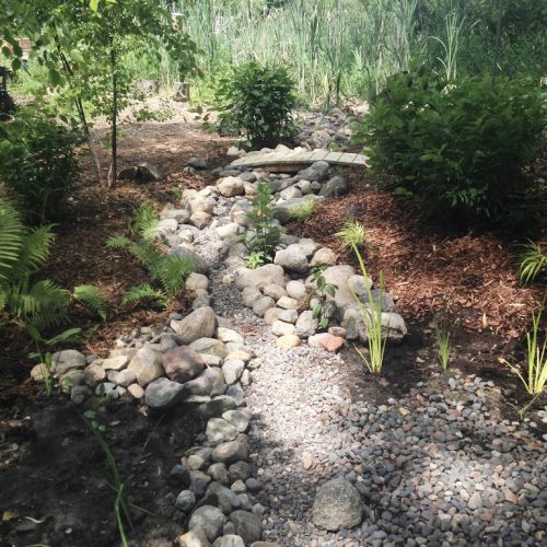 A rocky dry steam with a bird feeder, grasses, and a small bridge over the creek.