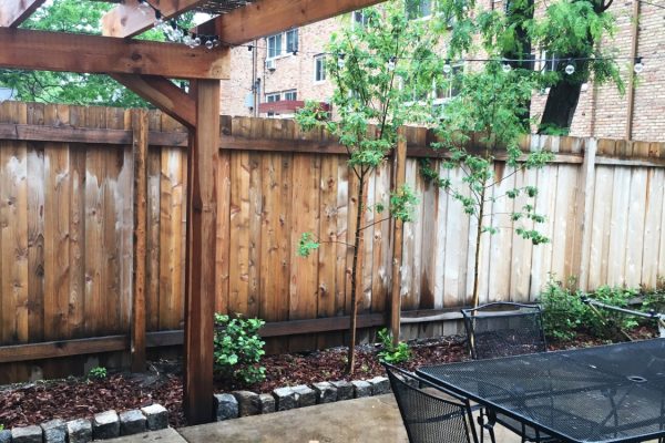 A wooden pergola covering a concrete patio with a black metal table and chairs in front of a wooden fence.