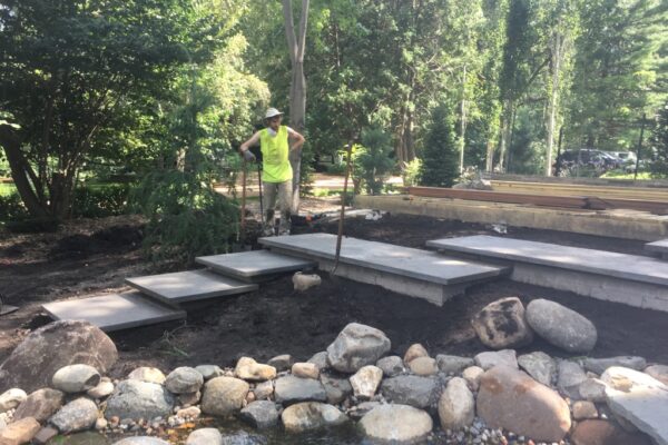 A construction worker standing behind a custom cascading stone staircase and water feature with large rocks in a lush wooded backyard.