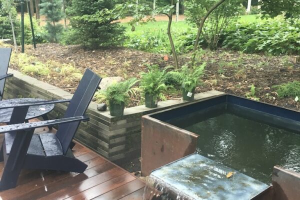 A modern custom metal cistern with flowing water on a wooden deck with black seating chairs, with a lush wooded lawn behind.