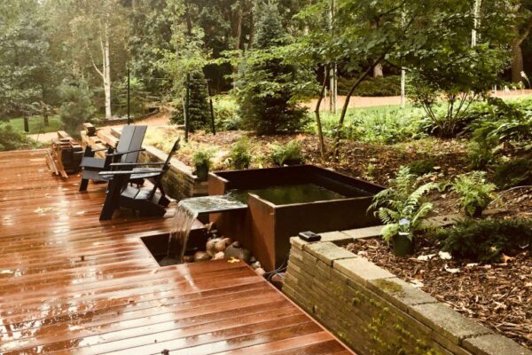 A wooden deck with a modern metal cistern with flowing water and with black seating chairs in front of a lush wooded lawn behind.