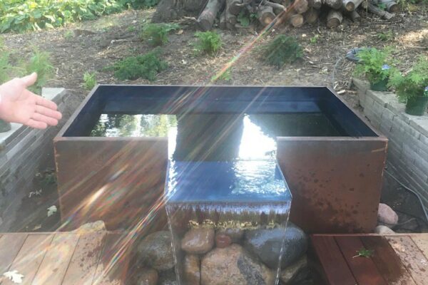 A square modern metal cistern with water flowing out of it onto rocks below, on a wooden deck.