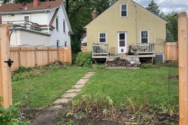 A short grassy lawn lined with shrubs in front of a yellow house with a wooden fence.