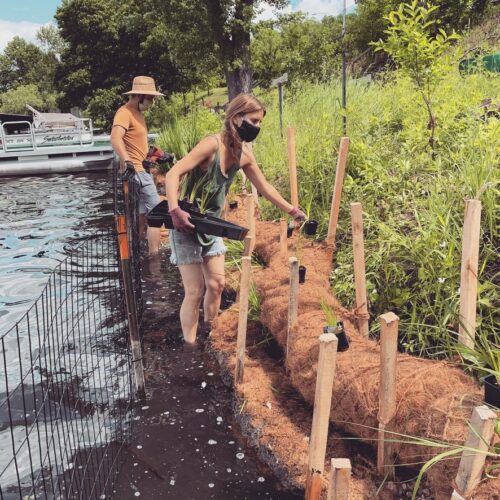 Two landscape crew members installing wooden stakes into coir logs from the water of Lake McCarrons against the sloped shoreline.