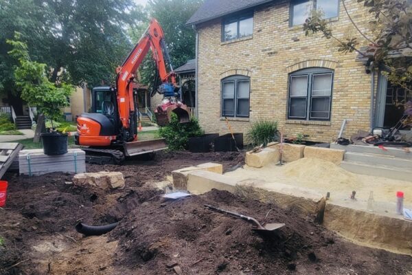 An orange mini excavator with grapple claw in motion prepares to pick up and move a 2'x4' mild steel bed that will be used for planting annual vegetables.