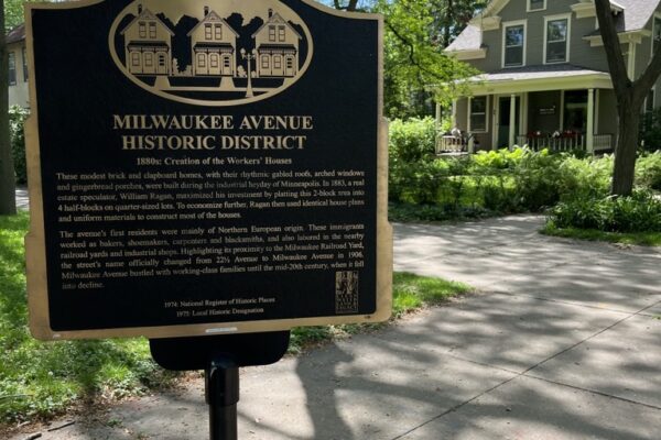 A black and gold illustrated sign explains the history of the Milwaukee Avenue Historic District and depicts an illustration of three Victorian era row houses.