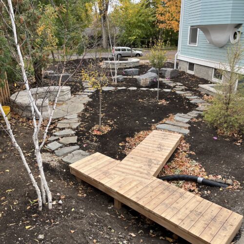 A sculptural, y-shaped wooden boardwalk connects to a rustic buff limestone paver path that encircles a newly planted woodland garden in the Kenwood Avenue neighborhood of Minneapolis.