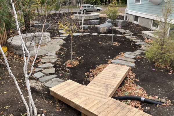 A sculptural, y-shaped wooden boardwalk connects to a rustic buff limestone paver path that encircles a newly planted woodland garden in the Kenwood Avenue neighborhood of Minneapolis.