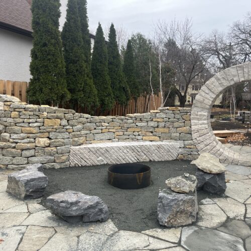 A flagstone patio, boulder-lined gravel fire pit, undulating dry-stacked wall and dramatic moon gate are key elements of a newly constructed outdoor kitchen and beer garden, situated on a private property on Kenwood Avenue in Minneapolis.