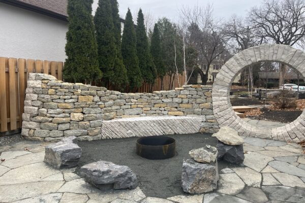 A flagstone patio, boulder-lined gravel fire pit, undulating dry-stacked wall and dramatic moon gate are key elements of a newly constructed outdoor kitchen and beer garden, situated on a private property on Kenwood Avenue in Minneapolis.