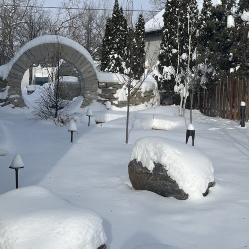 A heavy snow highlights the large rustic boulders, deciduous tree trunks and dramatic moon gate in a small and serene woodland garden in the Kenwood Avenue neighborhood of Minneapolis.