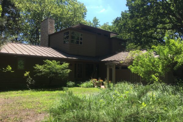 A stately prairie style house sits behind a large front yard with a patchy lawn surrounded by overgrown shrubs and messy grasses.