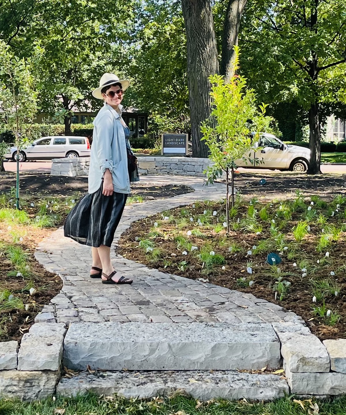 A woman wearing a hat, blue shirt, and black skirt standing on a cobbled stone path with new plants on either side on a tree-lined street for a portrait about her.