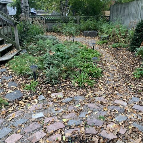 A rustic cobblestone fire pit connected to a circular seating area via a winding gravel path in a small shaded backyard in the city of Minneapolis.