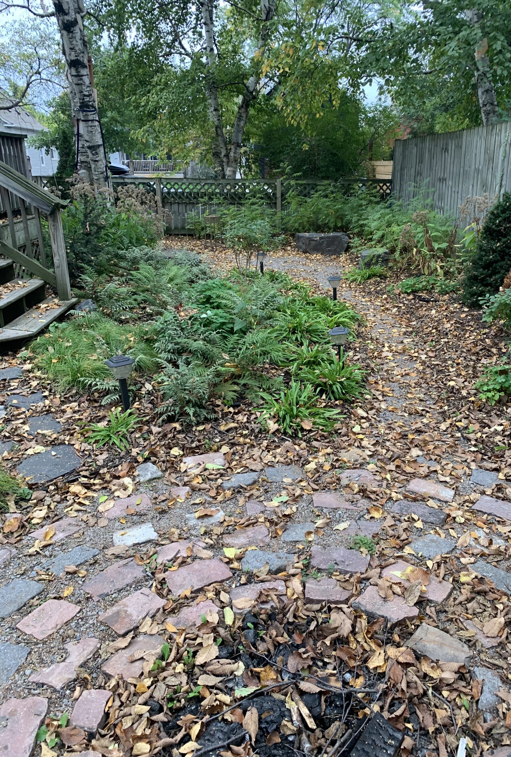 A rustic cobblestone fire pit connected to a circular seating area via a winding gravel path in a small shaded backyard in the city of Minneapolis.