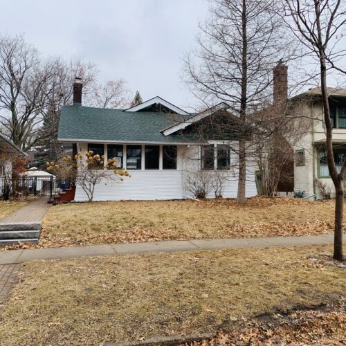 A quaint, cabin-like white house with green trim sits on a small lot with a bare lawn, foundation shrubs