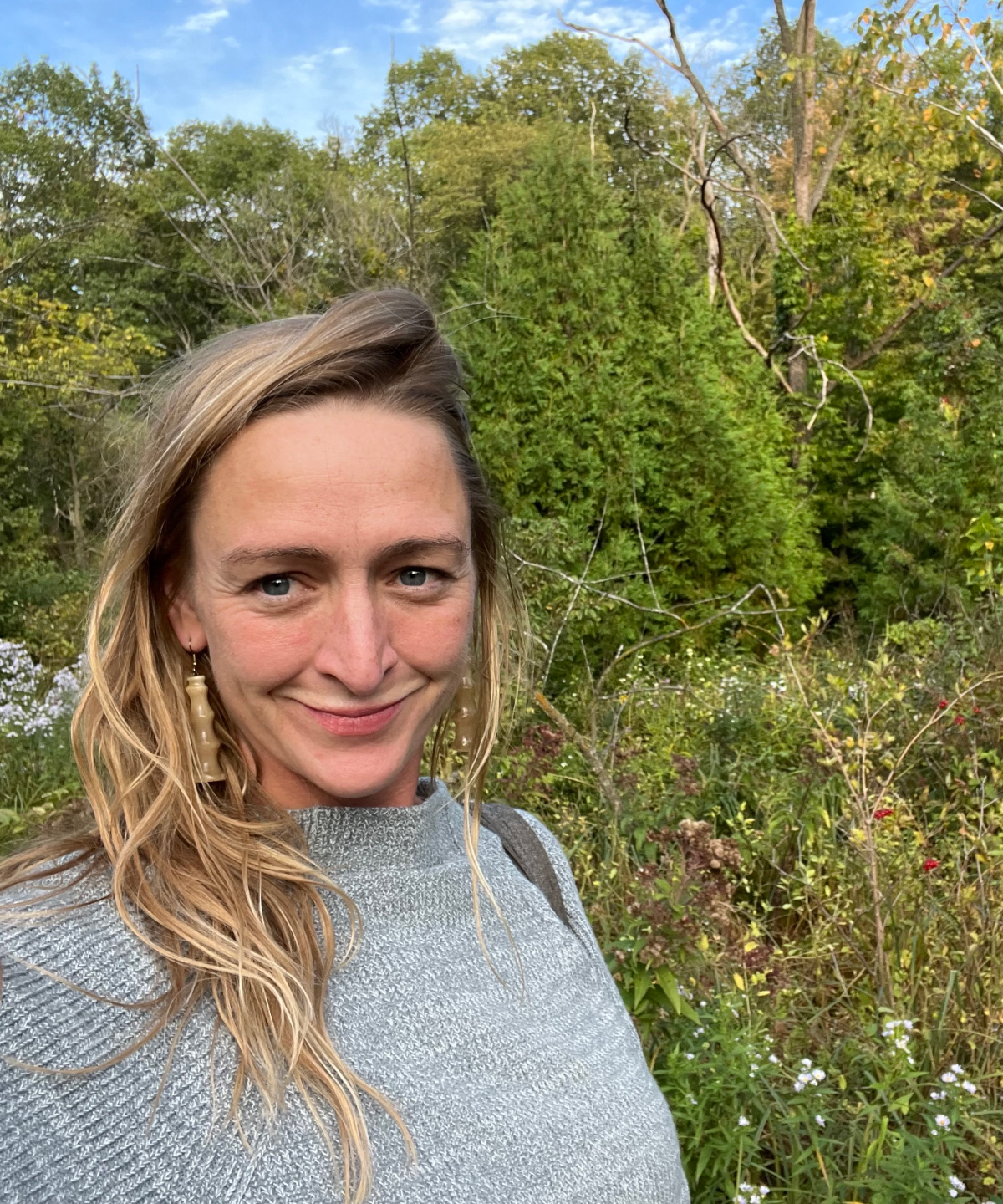 A portrait of a woman with blond hair smiling at the camera in front of a flowering tree filled landscape for a portrait about her.