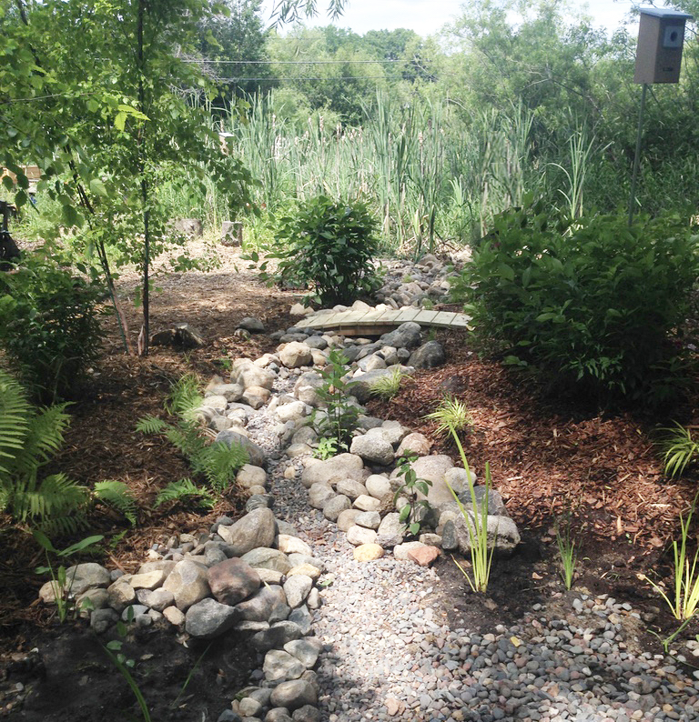 A rocky dry steam with a bird feeder, grasses, and a small bridge over the creek.