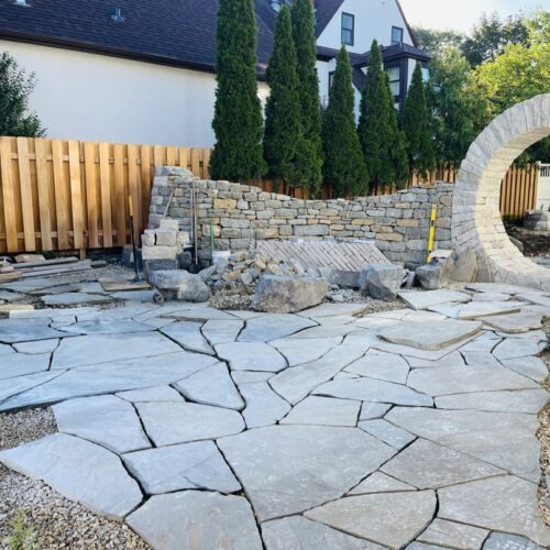 An in-progress flagstone patio, dry stacked rock wall and dramatic circular moon gate are key features of a rustic outdoor kitchen and beer garden.