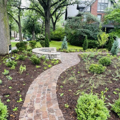 A newly constructed cobblestone path meanders through a freshly planted prairie garden toward a semi-circular stone seating circle, situated in a private front yard in the traditional and affluent Kenwood neighborhood of Minneapolis.